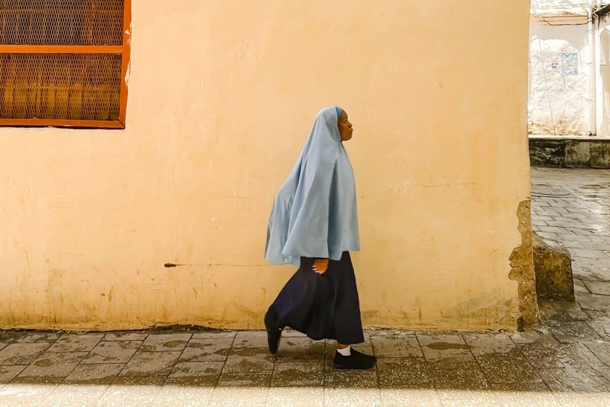 A woman in a light blue headscarf walks along a street past a sunlit wall in Iraq