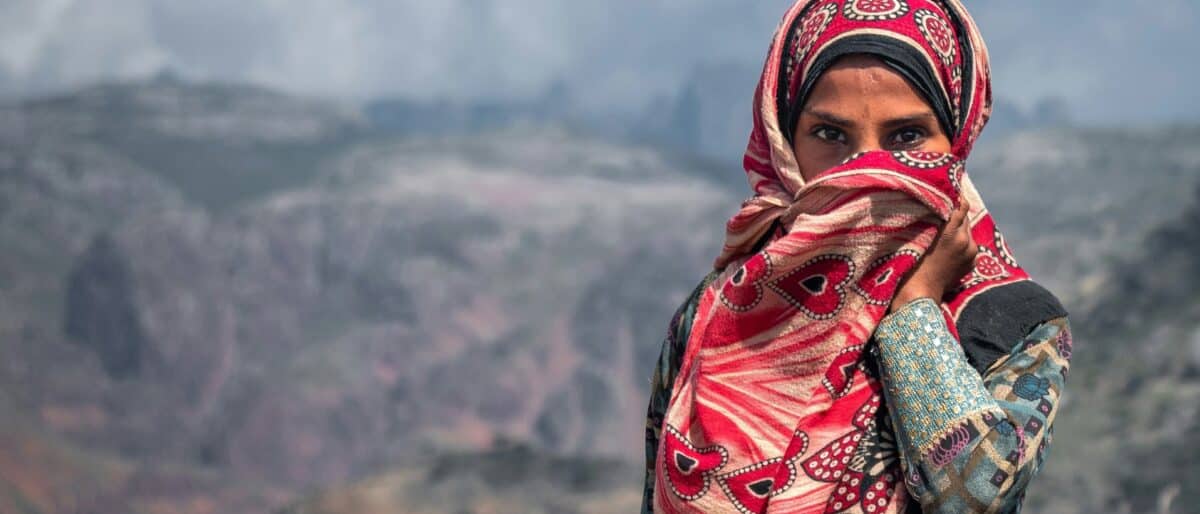 A woman in a red patterned headscarf stands before a mountain landscape in Yemen
