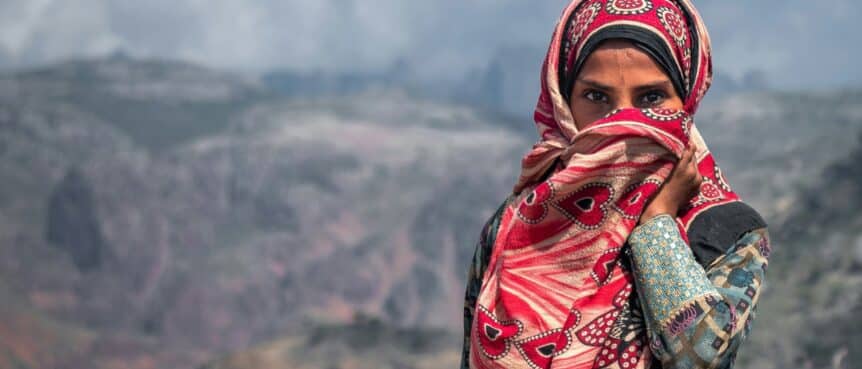 A woman in a red patterned headscarf stands before a mountain landscape in Yemen